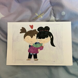 May include: A hand-drawn illustration on a white card depicts a couple embracing. The man, with brown hair, carries the woman, who has black hair, with a red heart above them. The woman wears a purple and green dress, and the man wears a blue shirt and black pants.