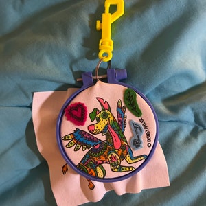 May include: A colorful embroidered design of a dog with wings, a heart, and other embellishments on a white fabric background. The design is in a blue embroidery hoop with a yellow plastic clip attached.