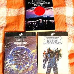 Könnte beinhalten: Drei polnische Taschenbücher im Vintage-Stil. Das obere Buch zeigt eine rote Sonne und den Titel "Dzień Czerwonego Giganta". Das mittlere Buch trägt den Titel "Taniec Potworów" mit einer blau-violetten Illustration. Das untere Buch trägt den Titel "Bogowie Naszej Planety" mit einer futuristischen Figur.