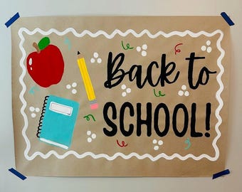 Puede incluir: Un letrero de papel marrón con las palabras "Back to SCHOOL!" en escritura negra. El letrero presenta una manzana roja, un lápiz amarillo y un cuaderno azul. El borde es blanco con un diseño ondulado. Cinta adhesiva azul asegura el letrero a la pared.