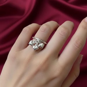 Könnte beinhalten: Ein silberner Ring mit drei herzförmigen Anhängern. Der Ring wird an einem Finger vor einem weinroten Hintergrund getragen. Das Design ist schlicht und elegant, passend für den Alltag oder besondere Anlässe.