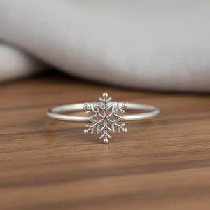 May include: A silver snowflake ring. The ring features a delicate, openwork snowflake design at the center. The band is thin and polished, and the snowflake has intricate details. The ring is set against a wooden surface.