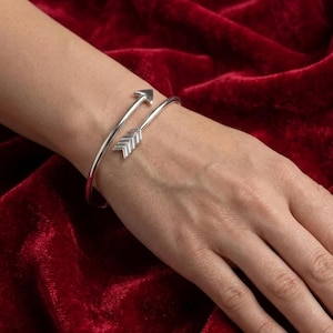 May include: A silver arrow cuff bracelet. The bracelet has an arrow design, with one end pointing towards the wrist and the other end pointing away. The bracelet is worn on a wrist.