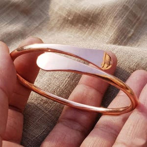 Puede incluir: Una pulsera de color cobre con un diseño único. La pulsera presenta una banda curva que se envuelve alrededor de la muñeca, con dos acentos planos en forma de hoja. El metal tiene una superficie pulida y reflectante, lo que le da un aspecto brillante. La pulsera es una joya.