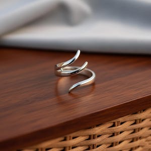 May include: A silver-colored ring with a unique, open design. The ring wraps around the finger with two curved, horn-like prongs extending upwards. The ring is displayed on a dark wooden surface.