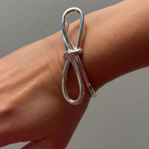 Puede incluir: Un brazalete plateado con un diseño en forma de lazo. El brazalete está hecho de un metal brillante y liso y presenta un detalle de nudo en el centro del lazo. El brazalete se lleva en la muñeca.