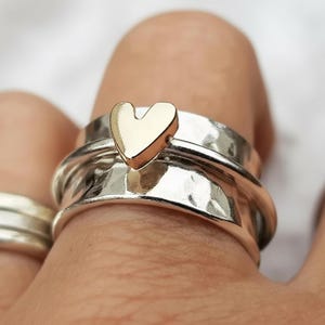 Può includere: Un anello d'argento con una texture martellata e un accento a forma di cuore dorato. L'anello è indossato su un dito, mostrando il contrasto tra il cuore dorato lucido e la fascia d'argento strutturata. È un gioiello.