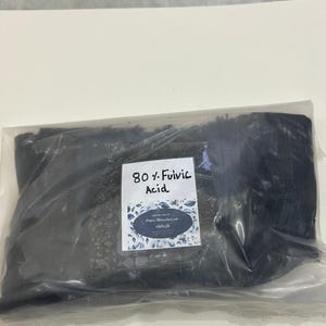 May include: A clear plastic bag filled with a dark, granular substance. A white label with the text "80% Fulvic Acid" and "Pure Himalayan Shilajit" is attached to the bag. The label has a floral design.