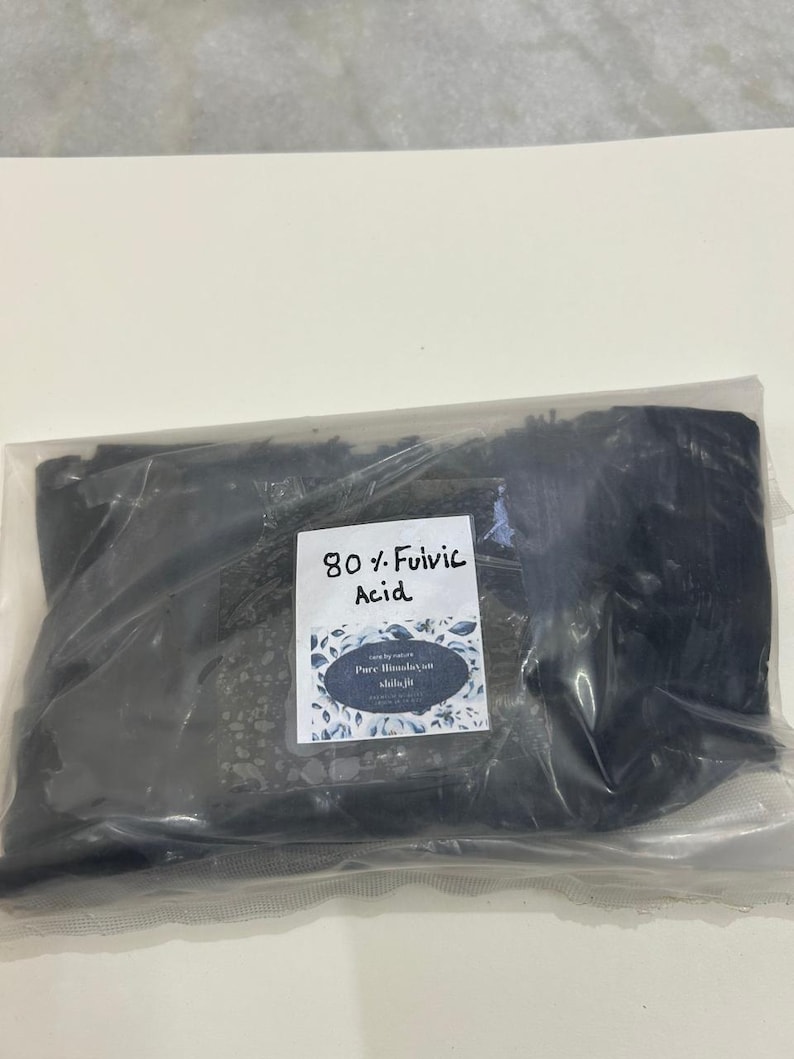 May include: A clear plastic bag filled with a dark, granular substance. A white label with the text "80% Fulvic Acid" and "Pure Himalayan Shilajit" is attached to the bag. The label has a floral design.