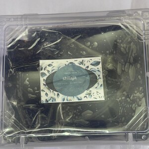 May include: A clear plastic container holding a dark, possibly organic substance. A small label with the word "shilajit" is visible, along with floral design elements. The container is sealed.