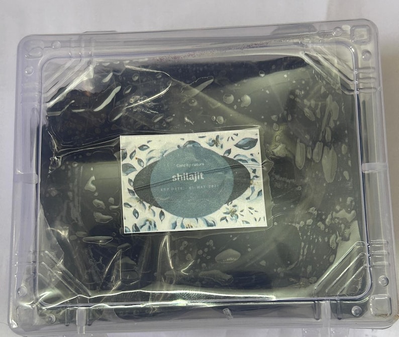 May include: A clear plastic container holding a dark, possibly organic substance. A small label with the word "shilajit" is visible, along with floral design elements. The container is sealed.