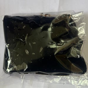 May include: A dark, rectangular object, possibly a food item, is encased in clear plastic. The object appears to be a solid, dark color, with a slightly glossy surface. The plastic packaging is crinkled, suggesting the item is soft or pliable.