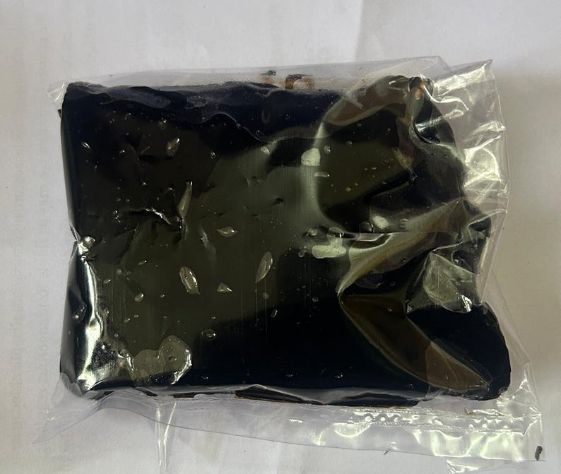 May include: A dark, rectangular object, possibly a food item, is encased in clear plastic. The object appears to be a solid, dark color, with a slightly glossy surface. The plastic packaging is crinkled, suggesting the item is soft or pliable.