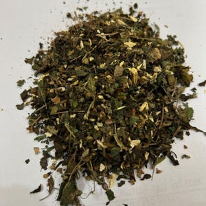 May include: A close-up of a pile of dried herbs and spices. The mix includes green leaves and light brown, flaky pieces. The blend suggests a variety of dried plants, potentially for cooking or medicinal purposes.
