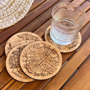 以下が含まれることがあります： ロンドン、ベルリン、パリなどの都市地図のデザインが施された木製コースター。透明なグラスがコースターの上に置かれ、他のコースターは木製のテーブルに重ねられています。コースターは丸く、詳細な通りのレイアウトが特徴です。