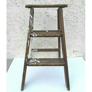 Könnte beinhalten: Ein Vintage-Trittschemel aus Holz mit drei Stufen und einer oberen Plattform. Der Hocker ist dunkelbraun gebeizt und hat weiße Farbspritzer. Der Trittschemel ist ein praktischer und dekorativer Gegenstand.