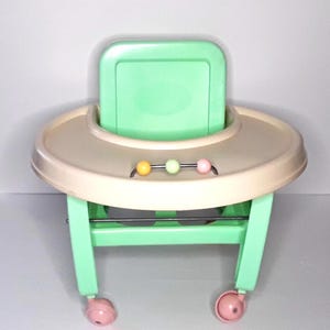 Peut inclure: Un trotteur pour bébé vert menthe et beige avec un plateau circulaire et un dossier. Le trotteur a un ensemble de trois perles colorées et des roues roses. Le cadre est vert menthe.