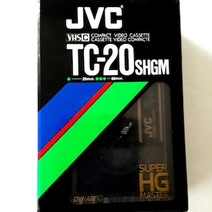 Könnte beinhalten: Schwarze JVC VHS-C Videokassette, Modell TC-20SHGM. Die Box hat ein diagonales Streifendesign in Blau, Grün und Rot. Das Band ist mit "SUPER HG MASTER" beschriftet und bietet eine Aufnahmezeit von 20 bis 60 Minuten.