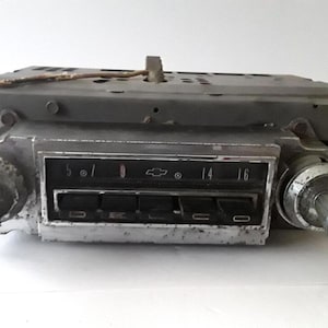 Könnte beinhalten: Vintage Autoradio mit silberner Blende und schwarzen Knöpfen. Das Radio hat ein Einstellrad mit Zahlen und ein Chevrolet-Logo. Das Metallgehäuse weist Alters- und Gebrauchsspuren auf.