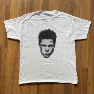 Könnte beinhalten: Weißes T-Shirt mit einem schwarz-weißen Porträt eines Gesichts auf der Vorderseite. Das Bild ist mittig platziert. Das T-Shirt scheint aus einem weichen, möglicherweise Baumwollstoff zu sein.