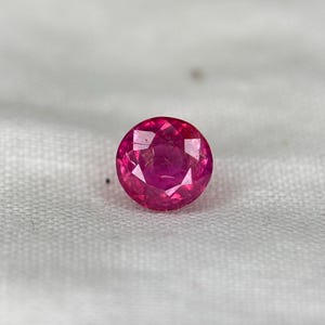 May include: A round, faceted ruby gemstone. The gem is a vibrant pinkish-red, with visible internal inclusions. The stone is set against a white, textured background, highlighting its colour and clarity. This gemstone is suitable for jewellery making.