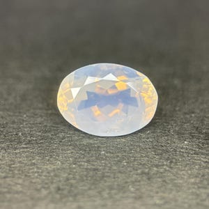 Op de afbeelding: Een ovaalvormige, gefacetteerde edelsteen met een melkachtig witte en goudgele interne gloed. De edelsteen is geplaatst tegen een donkergrijze achtergrond, wat de helderheid en glans benadrukt. Geschikt voor het maken van sieraden.