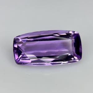 May include: A rectangular, faceted amethyst gemstone. The gem is a deep purple colour with a polished surface. The facets create a play of light and shadow, enhancing the stone's brilliance. The gemstone is set against a white background.