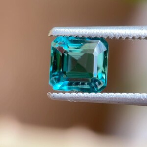 May include: A teal-coloured, square-cut gemstone is held by silver tweezers. The gemstone has a rectangular shape with multiple facets, reflecting light. The background is a blurred mix of brown and green.
