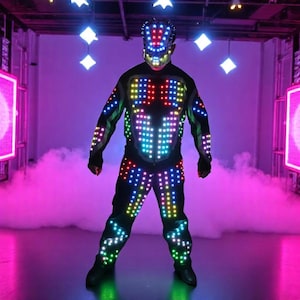 Op de afbeelding: Een zwart LED-lichtkostuum met bijpassend masker. Het pak is bedekt met veelkleurige lichten, gerangschikt in patronen over de borst, armen en benen. De achtergrond is een podium met roze en paarse verlichting en een rookmachine.
