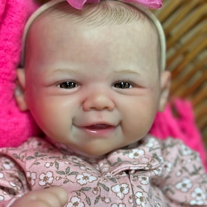 May include: A smiling baby doll with a pink floral onesie and a pink bow headband. The doll has fair skin, brown eyes, and a gentle expression. The background includes a pink blanket and a woven basket.