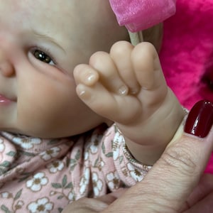 May include: Close-up of a baby doll's face and hand. The doll has brown eyes, fair skin, and a pink bow headband. The doll is wearing a pink floral print shirt. A hand with a burgundy nail polish is holding the doll's hand.