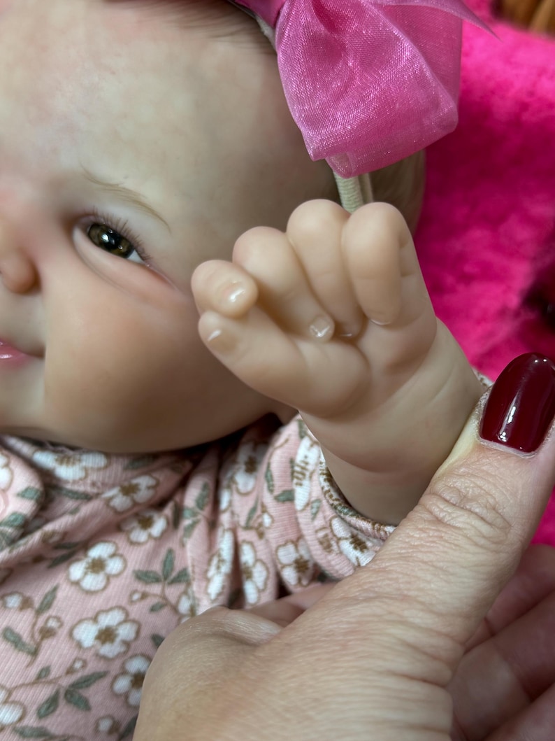 May include: Close-up of a baby doll's face and hand. The doll has brown eyes, fair skin, and a pink bow headband. The doll is wearing a pink floral print shirt. A hand with a burgundy nail polish is holding the doll's hand.