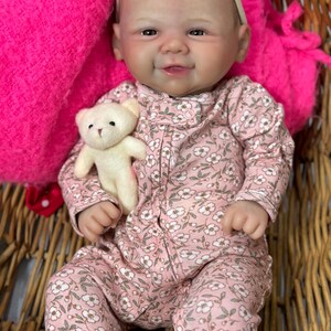 May include: A lifelike baby doll wearing a pink floral onesie and a matching headband with a pink bow. The doll is holding a small white teddy bear. The doll is in a wicker basket with a bright pink blanket.