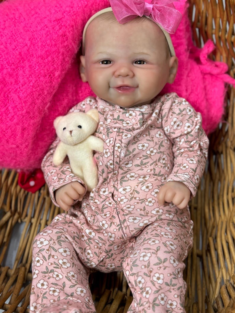 May include: A lifelike baby doll wearing a pink floral onesie and a matching headband with a pink bow. The doll is holding a small white teddy bear. The doll is in a wicker basket with a bright pink blanket.