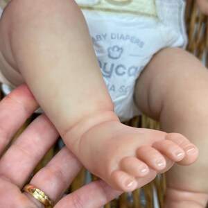 May include: Close-up of a baby's foot and leg, with a diaper visible. The skin is a pale pink color. The foot has tiny toes. The baby is in a woven basket.