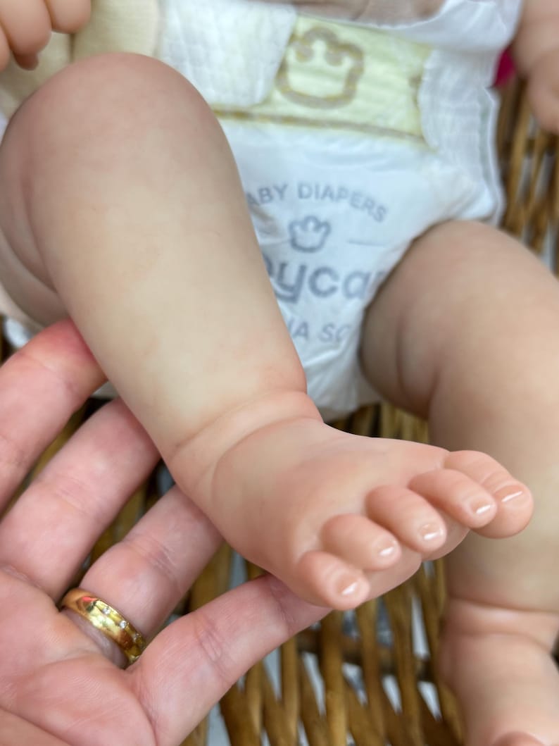 May include: Close-up of a baby's foot and leg, with a diaper visible. The skin is a pale pink color. The foot has tiny toes. The baby is in a woven basket.