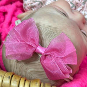 May include: A close-up of a baby's head with a bright pink bow. The bow is made of sheer fabric and is attached to a white headband. The baby is lying on a bed of pink fluff.