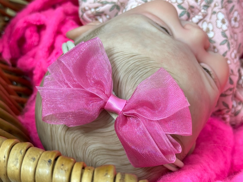May include: A close-up of a baby's head with a bright pink bow. The bow is made of sheer fabric and is attached to a white headband. The baby is lying on a bed of pink fluff.
