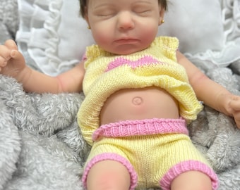 May include: A realistic baby doll with closed eyes and brown hair, wearing a pink headband with a bow. The doll is dressed in a yellow knitted top with a pink heart design and matching yellow shorts with pink trim, resting on a soft gray surface.
