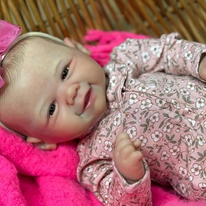 May include: A lifelike baby doll with a pink floral patterned onesie and a matching pink bow headband. The doll is lying on a bright pink blanket. The doll's skin has a realistic tone, and it has a gentle smile.