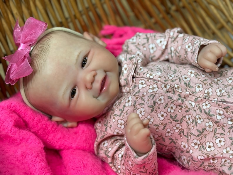 May include: A lifelike baby doll with a pink floral patterned onesie and a matching pink bow headband. The doll is lying on a bright pink blanket. The doll's skin has a realistic tone, and it has a gentle smile.
