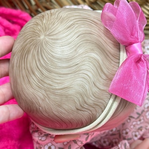 May include: A close-up of a doll's head with light brown hair styled in soft waves. A pink bow is attached to a cream-colored headband. The doll is wearing a floral patterned outfit.