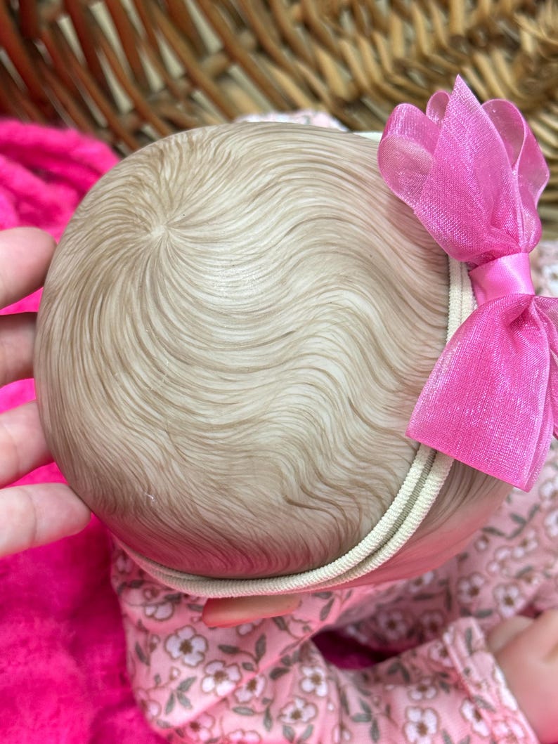 May include: A close-up of a doll's head with light brown hair styled in soft waves. A pink bow is attached to a cream-colored headband. The doll is wearing a floral patterned outfit.