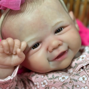 May include: A smiling baby doll with brown eyes and a pink bow. The doll is wearing a pink floral patterned outfit. The doll's skin has a realistic tone, and the details are finely crafted.