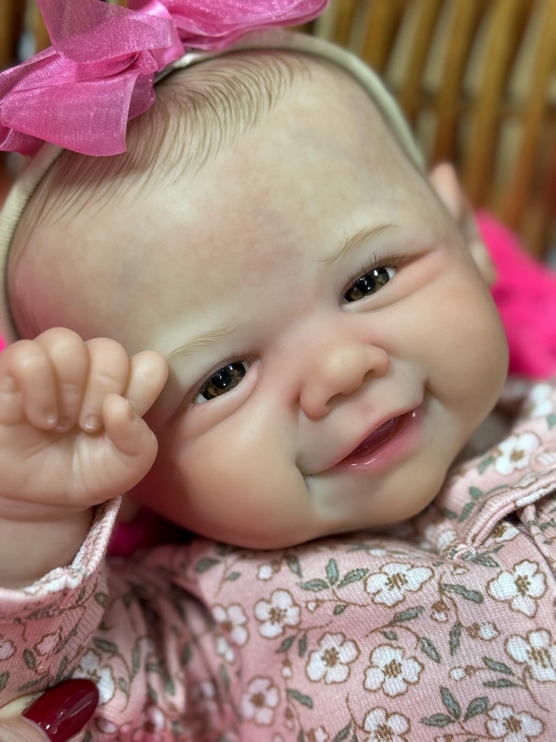 May include: A smiling baby doll with brown eyes and a pink bow. The doll is wearing a pink floral patterned outfit. The doll's skin has a realistic tone, and the details are finely crafted.