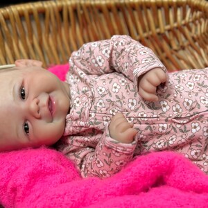 May include: A realistic baby doll with a smiling expression, wearing a pink floral patterned onesie and a white headband with a pink bow. The doll is lying on a bright pink blanket inside a woven basket.