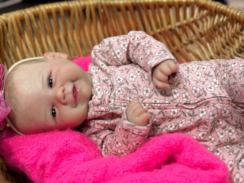 May include: A realistic baby doll with a smiling expression, wearing a pink floral patterned onesie and a white headband with a pink bow. The doll is lying on a bright pink blanket inside a woven basket.