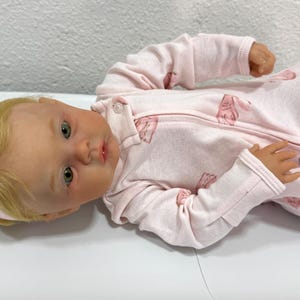 May include: A lifelike baby doll with blonde hair and green eyes, wearing a pink onesie with a zipper and a pink bow headband. The onesie features a repeating pattern of pink animal shapes.
