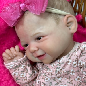 May include: A lifelike baby doll with a pink floral print onesie and a matching pink bow headband. The doll has realistic features, including detailed skin tones and brown eyes. The doll is lying on a pink blanket.