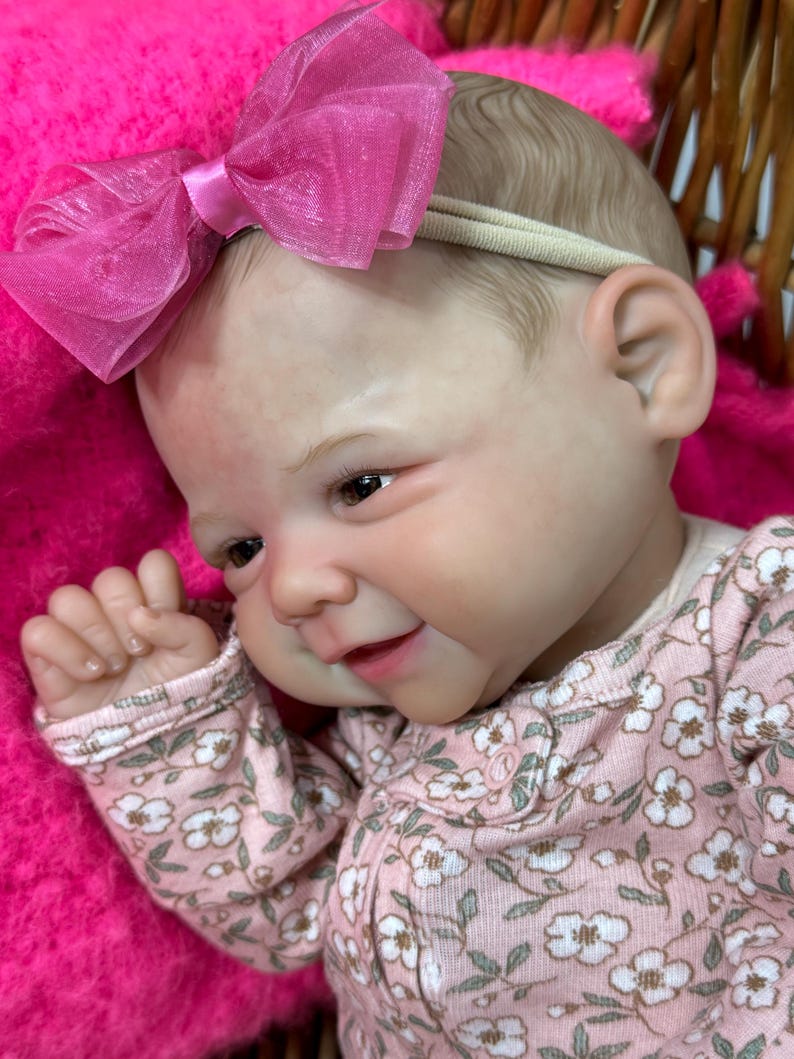 May include: A lifelike baby doll with a pink floral print onesie and a matching pink bow headband. The doll has realistic features, including detailed skin tones and brown eyes. The doll is lying on a pink blanket.